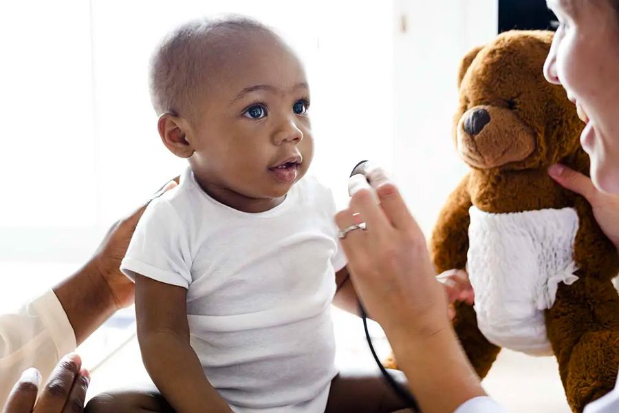 Individual Health Insurance - Baby during a Doctor Visit for a Wellness Checkup with the Doctor Holding a Child’s Teddy Bear to Try and Keep Child’s Attention and to Stay Positive