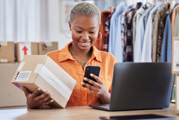 Blog - Woman Looking at Her Phone and Open Laptop While Organizing Packages