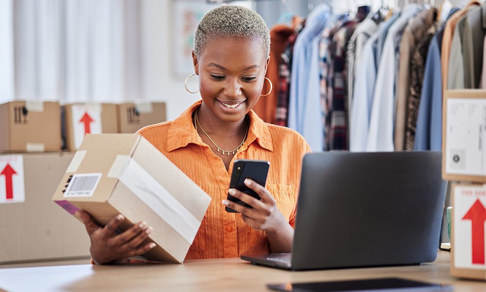 Blog - Woman Looking at Her Phone and Open Laptop While Organizing Packages
