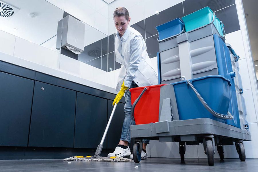 Janitorial Services Insurance - Female Janitor Mopping the Floor of a Restroom With Cleaning Cart Focus and View of Sinks in the Background